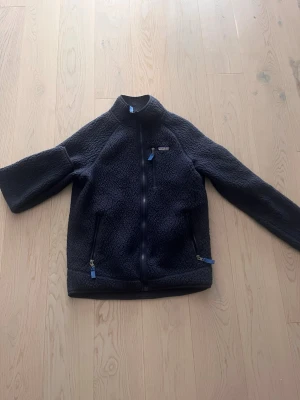 Svart fleecejacka från Patagonia - Mysig mörk blå fleecejacka från Patagonia med hel dragkedja och två fickor framtill med blå detaljer på dragkedjorna. Jackan har hög krage och är tillverkad i mjukt fleecematerial, perfekt för kyliga dagar. Pris kan diskuteras. 