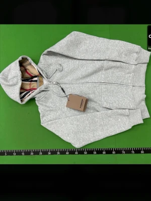 Burberry hoddie - Snygg gråmelerad hoodie från Burberry med dragkedja och klassiskt rutigt foder i huvan. Tröjan har två fickor fram, ribbade muddar och justerbar huva med snören. Perfekt för en chill och stilren look. Passar också för xs det står m men det stämmer inte