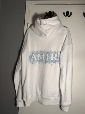 Vit AMIRI hoodie med blått tryck - Säljer en vit hoodie från AMIRI med blått prickigt tryck på bröstet och ryggen. Hoodien har en stor huva, magficka och ribbade muddar. Perfekt för dig som gillar streetwear och vill ha något snyggt och bekvämt.