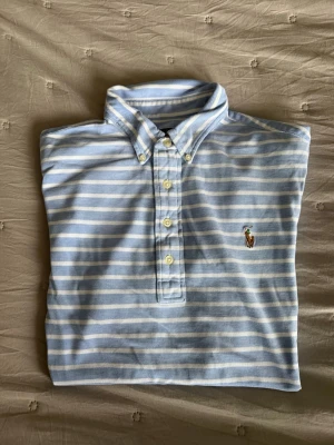 Blårandig skjorta från Ralph Lauren M strl - Snygg blårandig skjorta från Ralph Lauren med klassisk krage och knappar framtill. Skjortan har broderad logga på bröstet och är tillverkad i bomull. Perfekt för en stilren look.