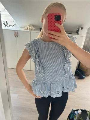 Blårandig blus med volangdetaljer - Söt blus med smala blå och vita ränder, volanger längs axlar och fram, samt broderade spetsdetaljer längst ner. Ärmlös modell med rund hals och peplum-kant. Perfekt för en fräsch och somrig look.