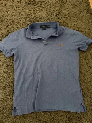 Blå pikétröja från Polo Ralph Lauren - Klassisk blå pikétröja från Polo Ralph Lauren med slim fit-passform. Tröjan har korta ärmar, krage och två knappar framtill samt den ikoniska broderade loggan på bröstet. Perfekt för en stilren och avslappnad look. Slim fit! 