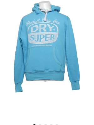 Ljusblå hoodie från Superdry  - Snygg ljusblå hoodie från Superdry med stort vitt tryck på bröstet. Klassisk känguruficka framtill, ribbade muddar och justerbar huva med vita snören. Perfekt för en avslappnad streetstyle-look.