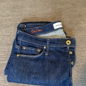 Slim-fit jeans från Jack & Jones - Mörkblå slim-fit jeans från Jack & Jones med klassisk femficksdesign och snygga guldfärgade knappar. Jeansen har raka ben och är tillverkade i ett slitstarkt denimtyg som passar perfekt till en avslappnad stil. Storlek 30, 32.