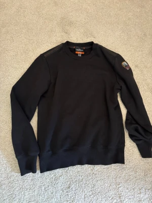 Svart sweatshirt från Parajumpers - Cool svart sweatshirt från Parajumpers med rund halsringning och ribbade muddar. Tröjan har en patch med logga på ena ärmen och detaljer på axlarna. Tillverkad i mjukt bomullsmaterial som är skönt att bära.