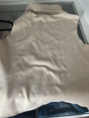 Beige stickad väst med polokrage - Snygg beige stickad väst med hög polokrage. Perfekt att styla över en skjorta eller t-shirt för en trendig lager-på-lager-look. Enkel och stilren design som passar till mycket.