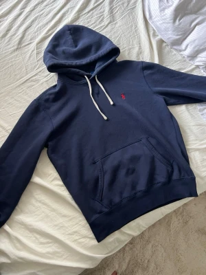 Ralph lauren hoodie - Mörkblå hoodie från Polo Ralph Lauren med vit dragsko i huvan och klassisk röd logga broderad på bröstet. Hoodien har en stor magficka och ribbade muddar vid ärmslut och nederkant. Perfekt för en avslappnad och stilren look.