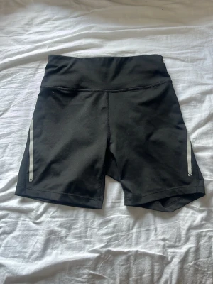 Svarta träningstights shorts från SOC - Svarta träningstights shorts från SOC med hög midja men kanten går att vika ner om man föredrar låg midja. Har reflex detaljer på sidorna och är i stretchigt material. Snygg och sportig design med tight passform.