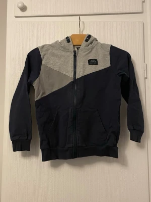 Mörkblå och grå hoodie med dragkedja - Snygg hoodie i mörkblått och grått med dragkedja framtill och två fickor. Hoodien har en cool färgblock-design och en huva i ljusgrått. Tillverkad i mjukt sweatshirtmaterial, perfekt för chill dagar.