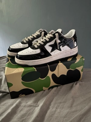 Svarta och vita BAPE Bapesta sneakers - Säljer ett par BAPE Bapesta sneakers i svart och vitt med glansig finish. Skorna har den ikoniska stjärnloggan på sidan, vita snören och detaljer i svart lack. Kommer med originalkartong i camomönster. Perfekta för dig som gillar streetwear och vill sticka ut. Priset är inte hugget i sten.