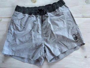Grå shorts från Sail Racing - Säljer ett par grå shorts från Sail Racing med svart resårmidja och snörning. Shortsen har en enkel design med logga på ena benet och meshfoder på insidan. Perfekta för sommardagar eller träning.
