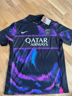 PSG x Nike svart matchtröja - Snygg svart PSG matchtröja från Nike med lila och blå grafiska detaljer. Tröjan har korta ärmar, rund hals och Paris Saint-Germain-logga på bröstet. Qatar Airways som sponsortryck på framsidan. Perfekt för fotboll eller streetwear.