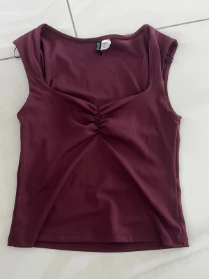 Vinröd ärmlös topp med rynk framtill - Snygg vinröd ärmlös topp med rynkdetalj vid bysten och djup, rundad urringning. Tillverkad i ett mjukt och stretchigt material som sitter tight mot kroppen. Perfekt att styla med jeans eller kjol för en trendig look.