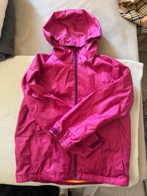 Rosa regnjacka Everest - Snygg rosa vindjacka med dragkedja framtill och praktisk huva. Jackan har långa ärmar med resår vid handlederna och är tillverkad i ett lätt, vindtätt material. Perfekt för blåsiga dagar och enkel att packa ner i väskan.