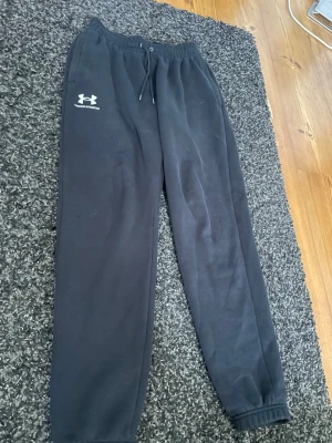 Svarta mjukisbyxor från Under Armour - Säljer ett par svarta mjukisbyxor från Under Armour med dragsko i midjan och resår vid bensluten. Byxorna har en klassisk passform och är tillverkade i mjukt material som känns skönt mot huden. Perfekta för chill eller träning. Köpt på jd sports 