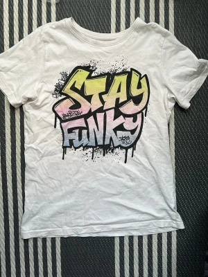 Vit t-shirt med graffiti-tryck - Vit t-shirt från Firefly med färgglatt graffiti-inspirerat tryck där det står 'Stay Funky' i gult, rosa och blått. Klassisk rund halsringning och korta ärmar. Perfekt för dig som gillar streetstyle och vill sticka ut.