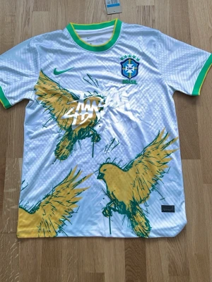 Vit Brasilien fotbollströja Nike och Stussy - Snygg vit Brasilien fotbollströja från Nike med gröna och gula detaljer. Framsidan har ett stort tryck med gula fåglar och texten 'stussy '. Tröjan har rund hals med grönt och gult kantband samt Brasilien-emblem på bröstet. Perfekt för match eller träning.