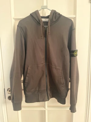 Grå zip hoodie från Stone Island - Säljer en grå hoodie från Stone Island med dragkedja framtill och två stora fickor. Klassisk huva med dragsko och den ikoniska Stone Island-loggan på vänster ärm. Tillverkad i mjukt bomullsmaterial, perfekt för en avslappnad streetwear-look. Tyvärr har den lite slitningar längst ner på vardera ärm (se bild) men annars bra skick.