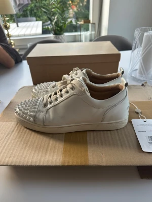 Christian Louboutin vita sneakers spikar  - Säljer ett par vita sneakers med coola nitar på tån och klassisk snörning. Skorna har en låg profil och är tillverkade i slätt läder med matchande vita sulor. Perfekta för dig som vill sticka ut med din stil.