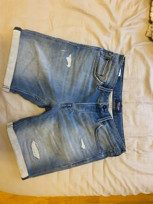 Blå jeansshorts från Jack & Jones - Säljer ett par blå jeansshorts från Jack & Jones med slitna detaljer och uppvikta benslut. Klassisk femficksmodell med knappgylf och normal passform. Perfekta för sommaren och riktigt snygga till en enkel t-shirt.