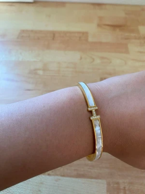 Guldigt armband med pärlemordetaljer - Snyggt armband i guldton med vita pärlemordetaljer längs hela bandet. Armbandet har en öppen design och ett H-format dekorativt spänne med små gnistrande stenar. Perfekt accessoar för att lyfta din stil. Köpt från Tunisien. Ej använd eftersom den är lite för stor för mig. 