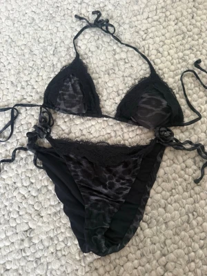 Svart leopardmönstrad bikini med spets - Snygg svart bikini med leopardmönster och spetsdetaljer. Överdelen är trekantsmodell med knyt i nacke och rygg, och underdelen har knyt i sidorna. Tyget hänger lite där bak på bikini trosorna, köpt för 2 år sen men bara använt den en sommar💕 