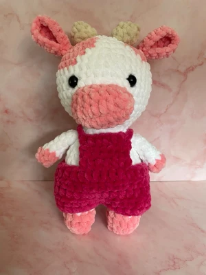 Mjukistjur i rosa hängselbyxor - Supergullig mjukistjur i vitt och rosa med horn och stora svarta ögon. Den har fluffig struktur och bär ett par stickade mörkrosa hängselbyxor. Perfekt som present eller dekoration i rummet.