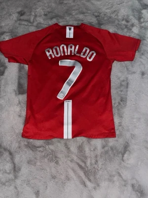 Manchester United Ronaldo matchtröja 2008 - Röd Manchester United matchtröja med vita detaljer, Ronaldo och nummer 7 på ryggen. Framtill finns klubbmärke, Nike-logga och AIG som sponsor. Tillverkad i funktionsmaterial, perfekt för fotboll eller samling.