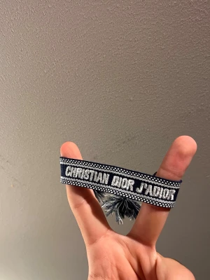 Christian Dior J'ADIOR blått armband - Säljer ett snyggt blått tygarmband från Christian Dior med texten 'J'ADIOR' i vitt. Armbandet har dekorativa tofsar i ändarna och ett vävt mönster längs kanterna. Perfekt accessoar för att lyfta din outfit.