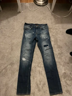 Blå slitna jeans med raka ben - Storlek 30/32         Säljer ett par blå jeans med raka ben och flera slitningar och hål på framsidan. Jeansen har klassisk femficksdesign och en tvättad look som ger en cool och avslappnad stil. Perfekta för dig som gillar streetwear och en trendig vibe. 