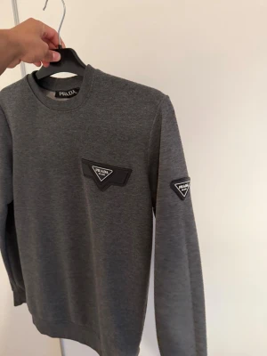 Grå sweatshirt från Prada - Snygg grå sweatshirt från Prada med klassisk rund halsringning. Tröjan har svarta Prada-loggor på bröstet och ena ärmen. Mjuk och bekväm passform, perfekt till jeans eller joggers.