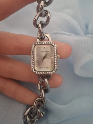 silverfärgad klocka med stenar - Snygg klockal i silverfärg med rektangulärt ur och glittrande stenar runt boetten. Kedjelänkad armbandsdesign och stilren urtavla med diskreta markeringar. Perfekt accessoar för dig som gillar lyxig och elegant stil.