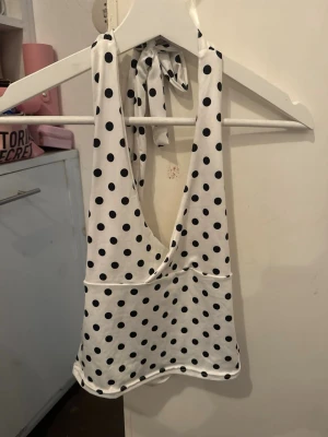 Vit prickig halterneck topp - Snygg vit halterneck topp med svarta prickar och djup v-ringning. Toppen är ärmlös och knyts i nacken för en trendig look. Perfekt till jeans eller kjol för en cool och lekfull stil.