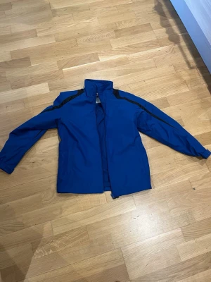 Blå south west vindjacka  - Snygg blå vindjacka med svarta detaljer längs axlar och ärmar. Jackan har hög krage, dragkedja framtill och justerbar nederkant. Perfekt för blåsiga dagar och enkel att matcha med olika outfits. Materialet känns lätt och smidigt.