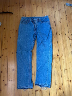 Blå raka jeans med rå kant - Säljer ett par klassiska blå jeans med rak passform och hög midja. Jeansen har fem fickor. Uppsprättat nere vid fötterna så dom är någon centimeter längre. Tillverkade i denim med tydliga sömmar och utan synligt märke. Köpta från Carlings och är deras egna märke Vailent. Storlek XS. 165 cm på bilden.
