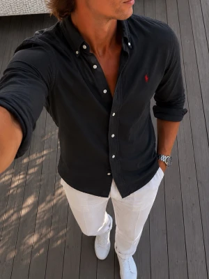 Svart Ralph Lauren skjorta med knappar - Snygg svart skjorta med klassisk krage och vita knappar. Skjortan har lång ärm och en liten röd broderad logga på bröstet. Perfekt till både jeans och chinos för en clean look.