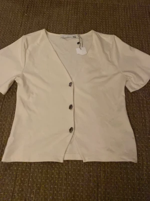 Vit kortärmad topp med knappar - Stilren vit topp med v-ringning och tre dekorativa knappar framtill. Kortärmad modell i mjukt material, perfekt för en enkel och clean look. Passar till både jeans och kjol för en avslappnad vibe.