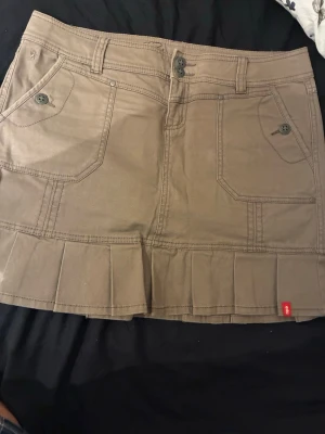 Beige kortkjol från Esprit - Säljer en beige kortkjol från Esprit med stora fickor framtill, knapp- och dragkedjestängning samt plisserad nederkant. Kjolen har bälteshällor och en casual look som passar perfekt till sommaren.