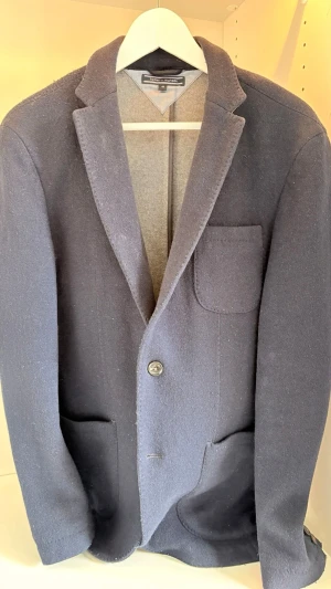 Mörkblå kavaj från Tommy Hilfiger - Stilren mörkblå kavaj från Tommy Hilfiger med klassisk passform. stickat material som ger en avslappnad men ändå dressad look. Perfekt för dig som vill ha en stlig kavaj nu till sommaren