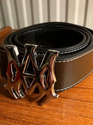 Svart bälte AMIRI - Snyggt svart bälte från AMIRI i skinn med vit söm längs kanterna och ett stort, unikt metallspänne i silverfärg. Perfekt för att ge outfiten en edgy touch. Bältet har en bred design och cool detalj med det iögonfallande spännet. HELT ny, kommer med hålgörare.