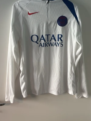 Psg tröja  - Säljer en vit långärmad fotbollströja från Paris Saint-Germain med blå detaljer och röd Nike-logga. Tröjan har tryck med Qatar Airways på framsidan och PARIS i blått på ryggen. Tillverkad i ett lätt och ventilerande material, perfekt för träning eller match.