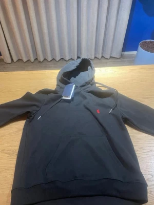 Svart hoodie från Polo Ralph Lauren - Snygg svart hoodie från Polo Ralph Lauren med klassisk röd logga broderad på bröstet. Hoodien har huva med dragsko och en stor magficka framtill. Tillverkad i mjukt material som känns skönt mot huden. Perfekt för en avslappnad och trendig stil. Priset är inte huggit i sten och kan diskuteras. HOODIEN ÄR M MEN PASSAR S. 