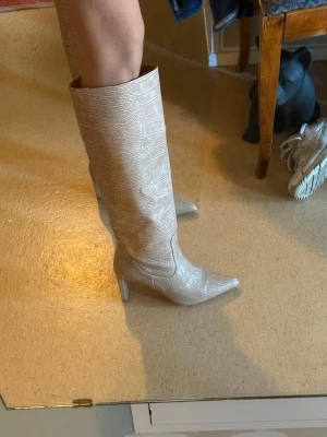 Beige krokopräglade höga stövlar - Säljer ett par beige höga stövlar med spetsig tå och blockklack. Stövlarna har ett trendigt krokodilpräglat mönster i konstläder och når upp till knäna. 7cm klack