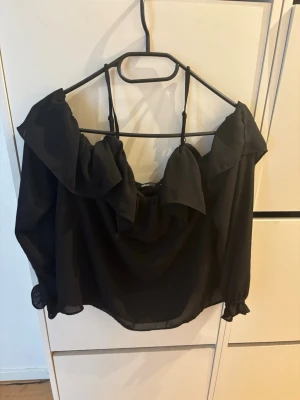 Svart offshoulder blus med volang - Snygg svart offshoulder blus med volangdetaljer runt halsringningen och tunna axelband. Blusen har långa ärmar med resår vid ärmslut och är gjord i ett lätt, transparent tyg som ger en luftig känsla.