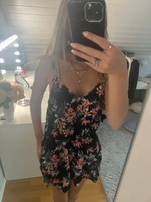 Svart blommig sommarklänning - Svart kortklänning (jumpsuit) med blommigt mönster i rosa och vitt. Klänningen har smala axelband och v-ringad halsringning. Perfekt för varma dagar och enkel att styla med smycken. Storlek 42 och sitter som en xs/s❤️