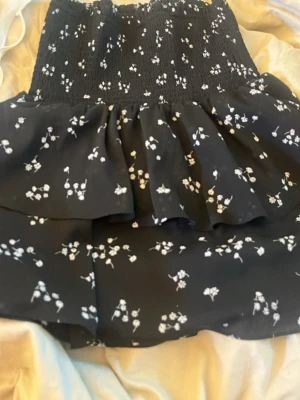 Svart blommig volangkjol - Söt svart kortkjol med vita små blommor och volangdetaljer. Kjolen har en smockad midja som ger en bekväm passform och ett luftigt fall. Perfekt för dig som gillar en feminin och trendig stil.