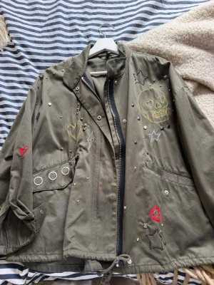 Olivgrön jacka med broderier - Unik olivgrön jacka med dragkedja och coola broderade detaljer. Köptes från HM för nått år sen men inte kommit till användning, den är som ny! Jackan har snörning i nederkant, stora fickor och dekorativa nitar. Oversized så skulle säga storlek 36 men passar 34.