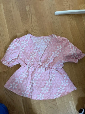 Rosa blommig blus med puffärm - Söt rosa blus med vita blommor och puffiga korta ärmar. Blusen har v-ringning och resår i midjan för en feminin siluett. Materialet är lätt och har struktur med broderade blommor som ger en romantisk känsla.