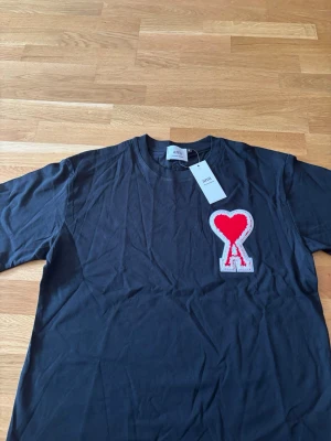 Svart t-shirt med rött hjärta från Ami - Svart t-shirt från Ami med ett stort rött hjärta och bokstaven A broderat på bröstet. Klassisk rund hals och korta ärmar. Tillverkad i mjuk bomull för en skön känsla. Perfekt för dig som gillar stilrena plagg med en twist.