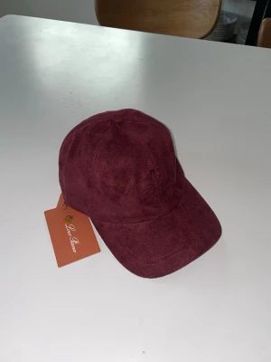 Vinröd keps Loro piana  - Snygg vinröd keps i mocka från Loropiana med justerbart spänne bak i läder. Kepsen har en klassisk böjd skärm och diskret broderad logga framtill. Insidan är fodrad med tyg och har en orange kant. Perfekt accessoar för en trendig look.